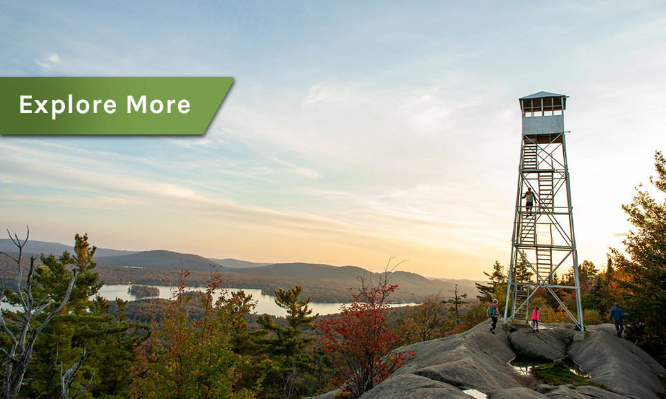 A family climbs and stands below an Adirondack fire tower on a mountain top, with the settling sun glowing over a forest of autumn foliage.